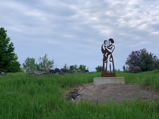 A sculpture of a woman holding a baby stands near the foundation ruin of a miner's house.