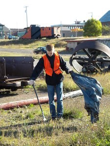 A youth volunteer uses a stick to pick up trash and put it in a trash bag outside.