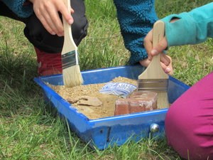 Hands use a brush to wipe sand away from artifacts.