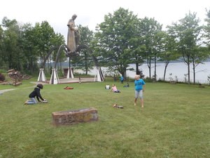 A group of children play in the grass next to a large, gold colored statue of a man holding a cross.