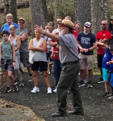 A park ranger in a gray shirt and green pants is talking to a large group of people.