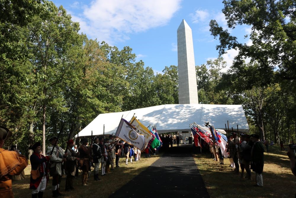 A large group of living historians lines up outside a tent. There is a large stone monument in the background surrounded by trees.