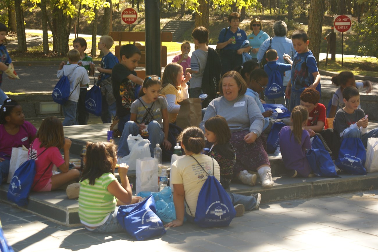 A large group of students are eating lunch with their teachers. Some are seated and some are standing near a concrete area.
