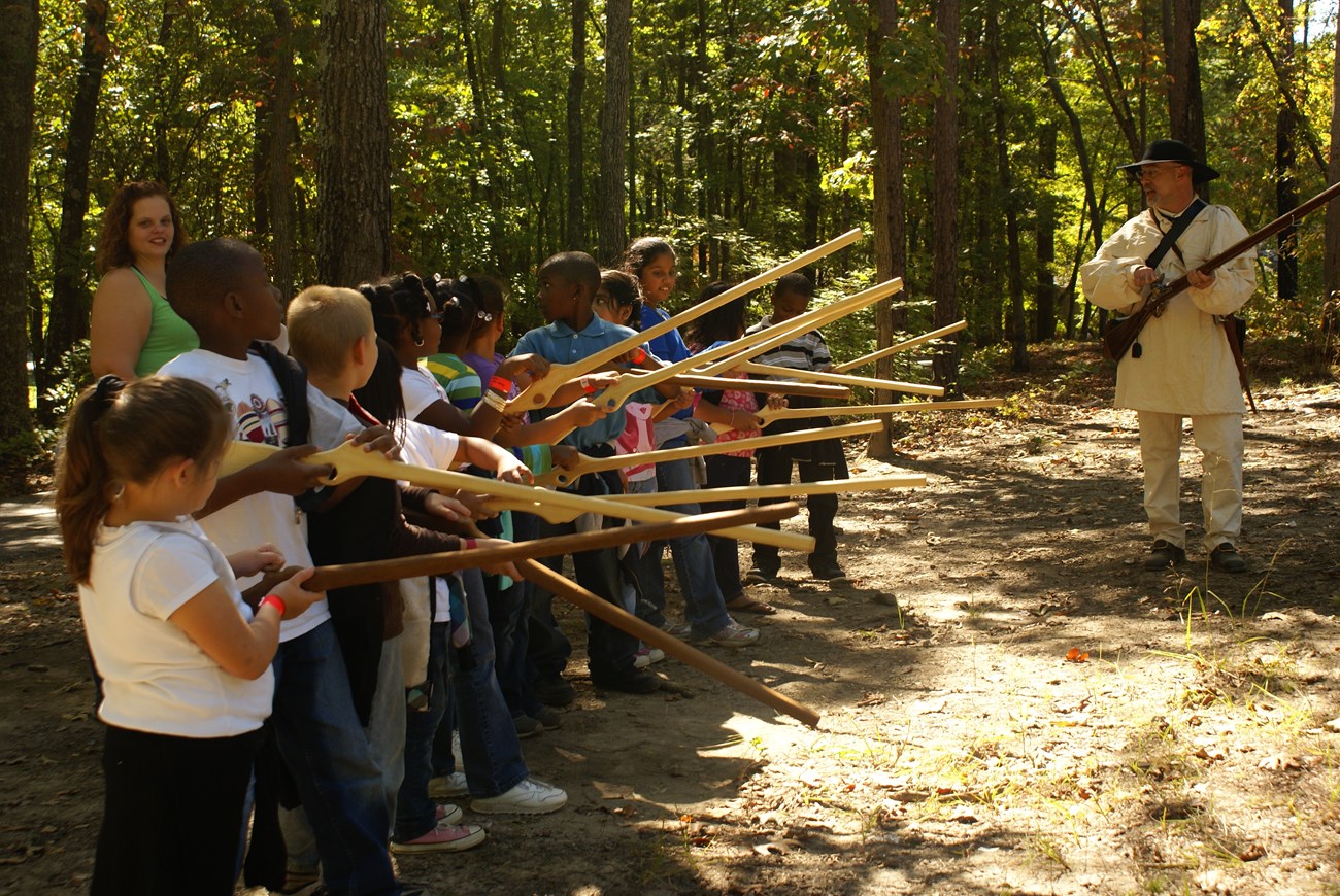 A living historian in a white hunting shirt and carrying a musket is talking to students and a teacher. The students are carrying wooden fake muskets. There are trees in the background.