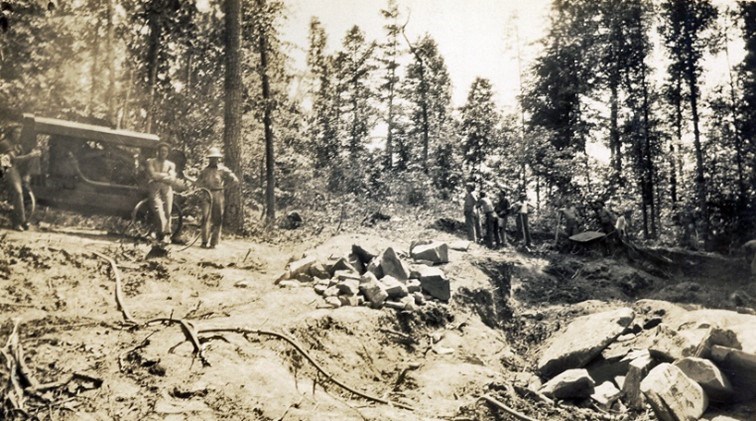 A black and white photo of two men near an antique car with woods and rocks around them.