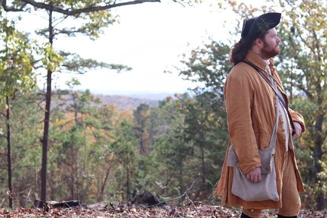 A man in a tan hunting coat looks to his left. He is surrounded by trees.
