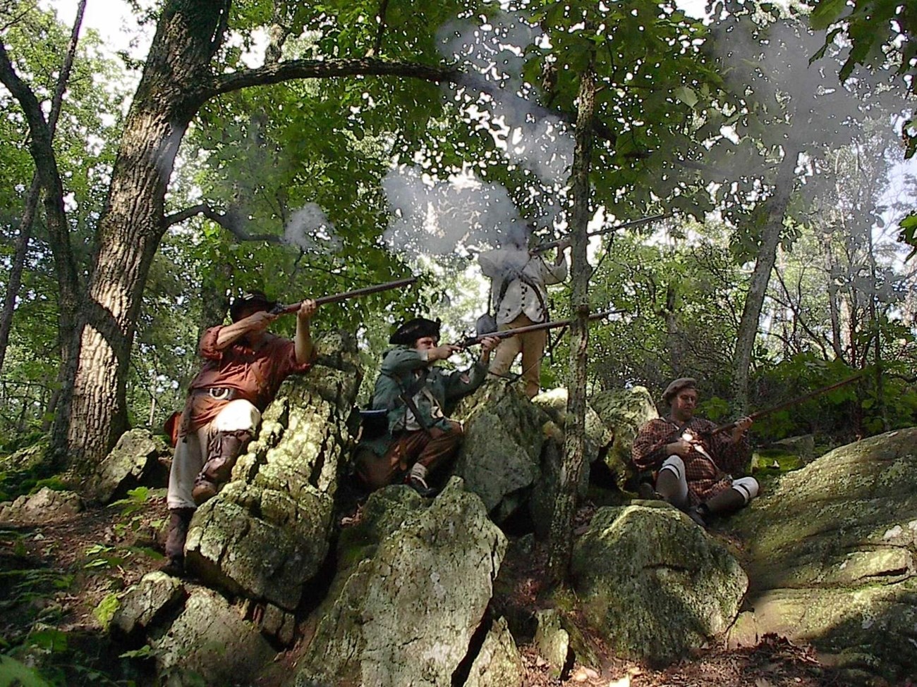 Men in 18th century uniforms on firing muskets while sitting and standing by rocks.