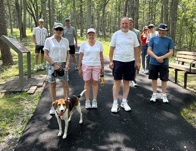 A group of 8 people and a dog stand along a paved trail surrounded by woods.