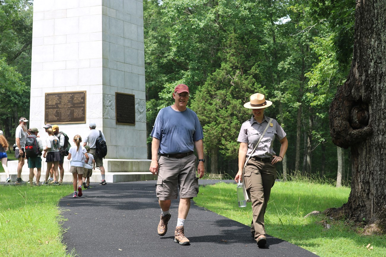A visitor and a park ranger walk a trail with a monument behind them.