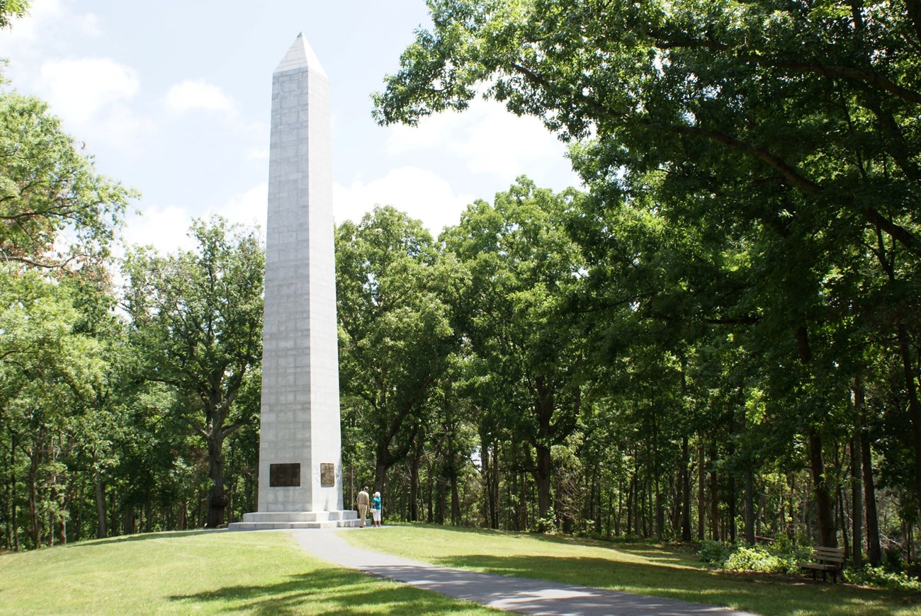 A large white monument is in the woods next to a paved hiking trail.