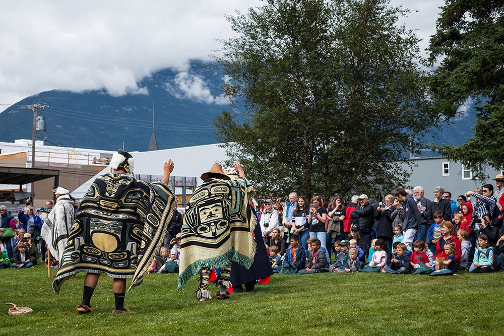 Two people in traditional Chilkat blankets dance in front of a large crowd