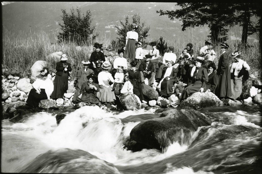 Black and white photo of women and children sitting by a flowing river in a natural setting.