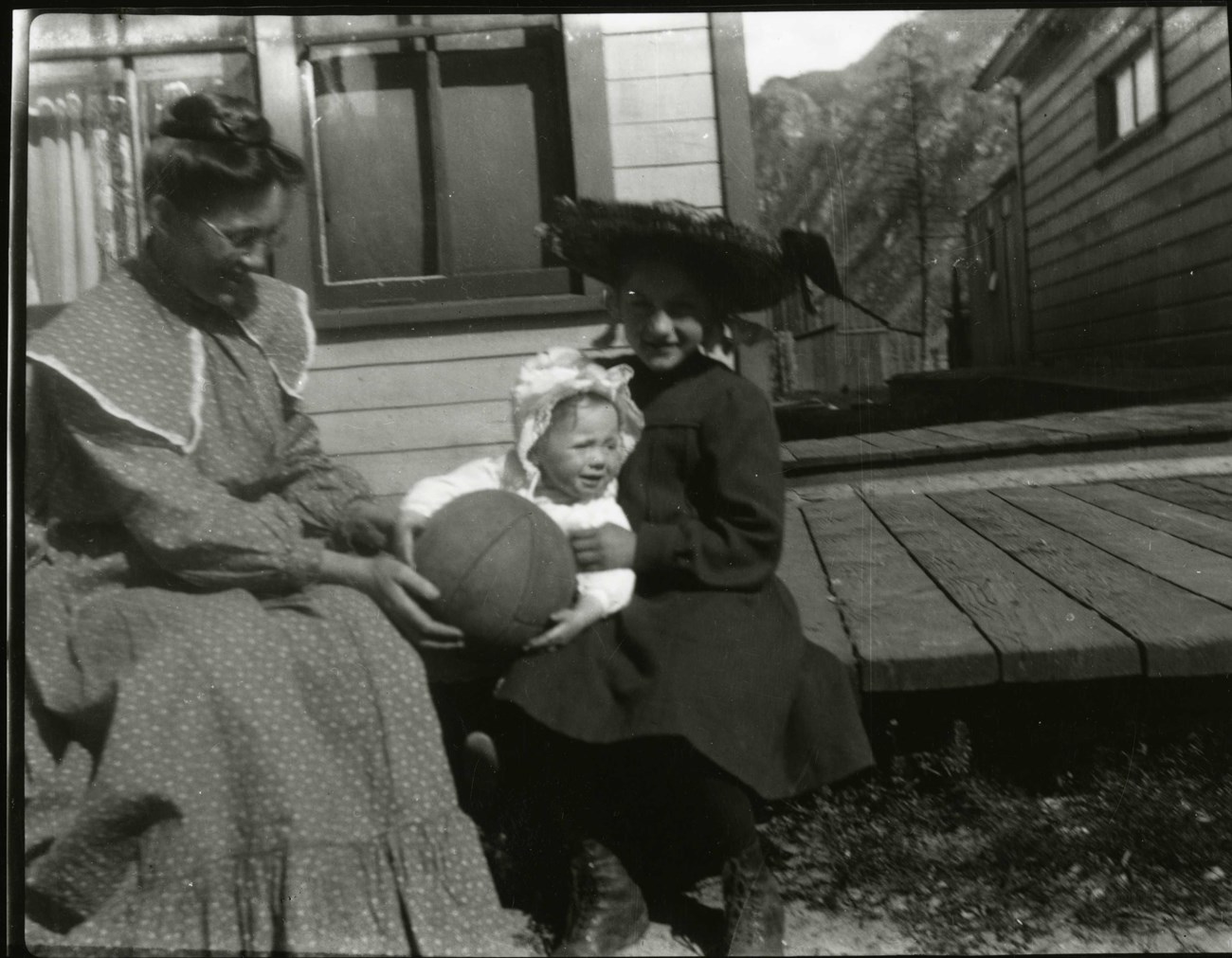 Black and white photo of two women sitting on a wooden platform playing with a baby who is holding a basketball.