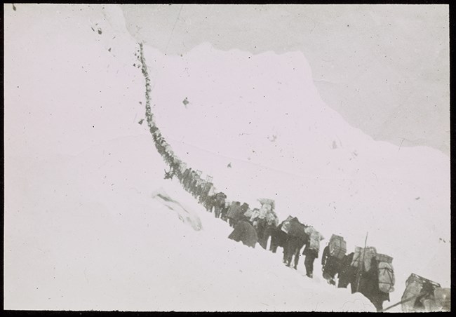 Black and white photo of line of people climbing a steep, snowy slope