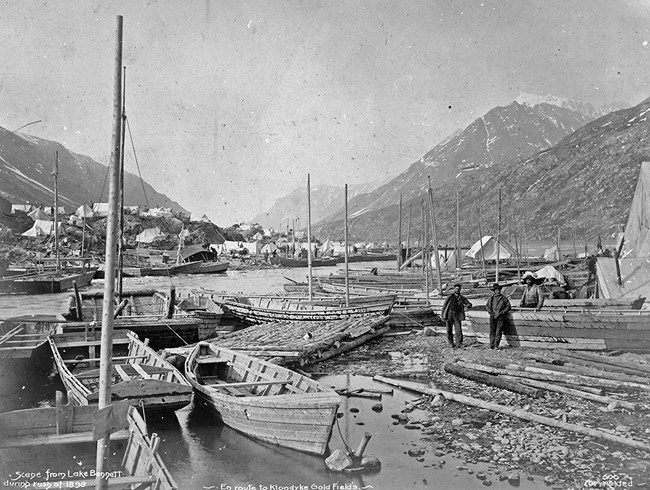 Black and white photo of rough wooden boats stretching from foreground to background. Two men pose midground with mountains and tent city behind them.