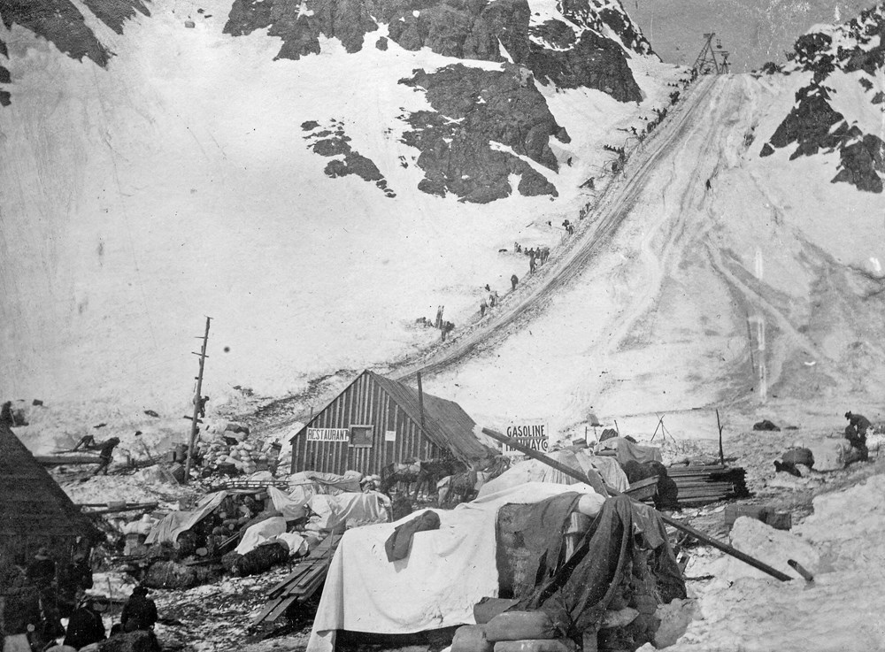 Historic photo of the line of people walking up steep snowy pass with buildings and supplies in foreground