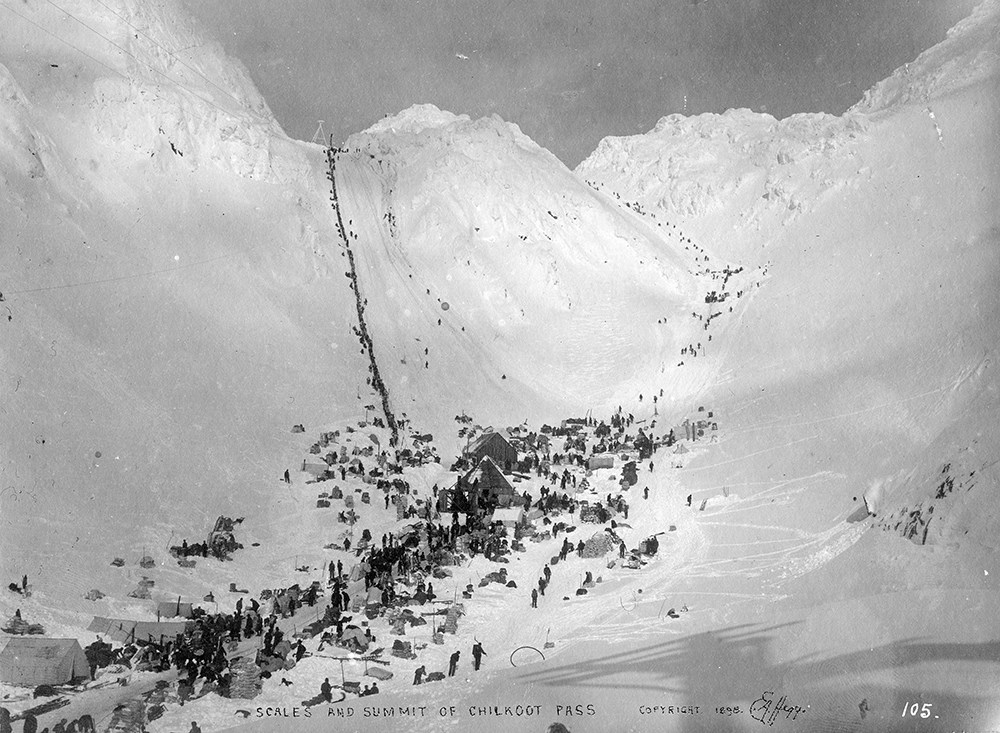 Black and white photo of tents and people collected at the base of two snowy mountain passes.
