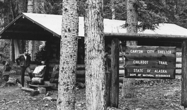 Black and white photo backpacker standing on porch of log cabin with a sign reading "Canyon City Shelter Chilkoot Trail State of Alaska Dept of Natural Resources"