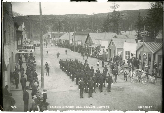 Citizens and wooden buildings line a dirt road on both sides. Soldiers line up in formation in the center of the street