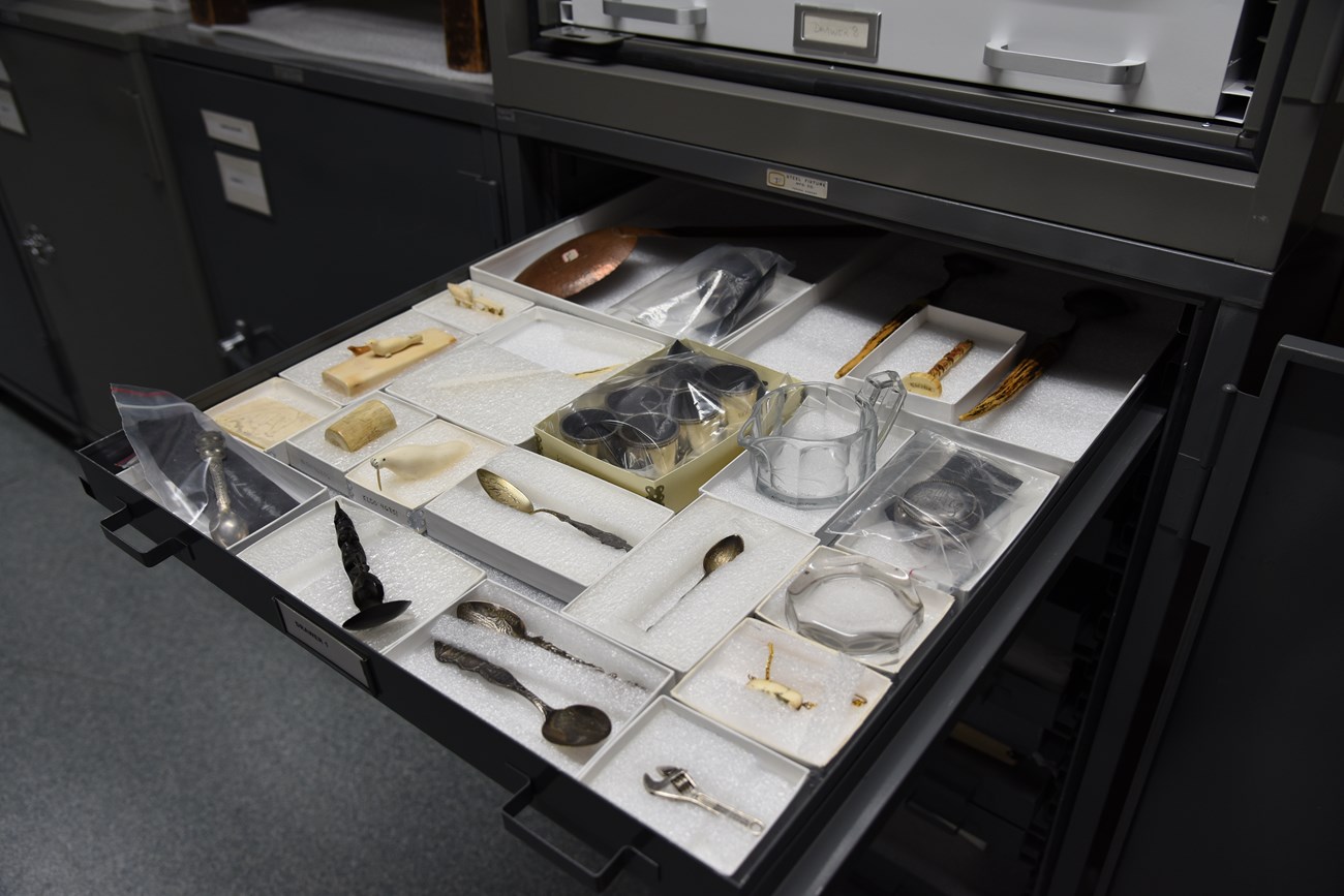 color photo of a wide and shallow grey drawer filled with small open topped white boxes housing various small artifacts