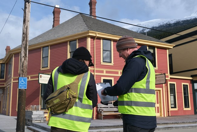 Photo of individuals wearing fluorescent vests and standing in front of the well-maintained historic Visitor Center in downtown Skagway while reviewing notes on a clipboard.