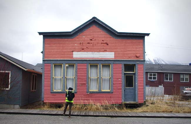 Photo of a person wearing fluorescent vest and taking notes in front of a Skagway historic building that is lifted on blocks, has flaking paint, and does not appear to be in use.