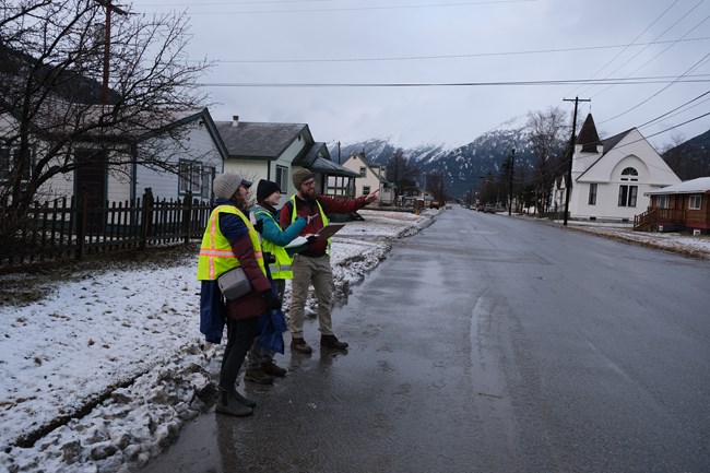 Photo of three individuals in neon yellow vests stand together as they observe a building on the other side of the street. There is an overcast sky and snow on the ground.