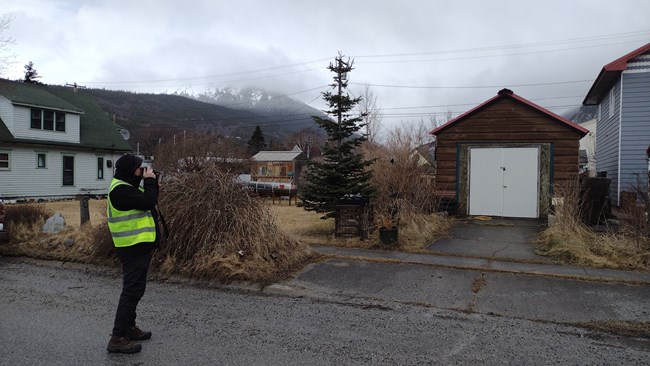 Photo of an individual in a neon yellow vest holding a camera and facing a house in Skagway.
