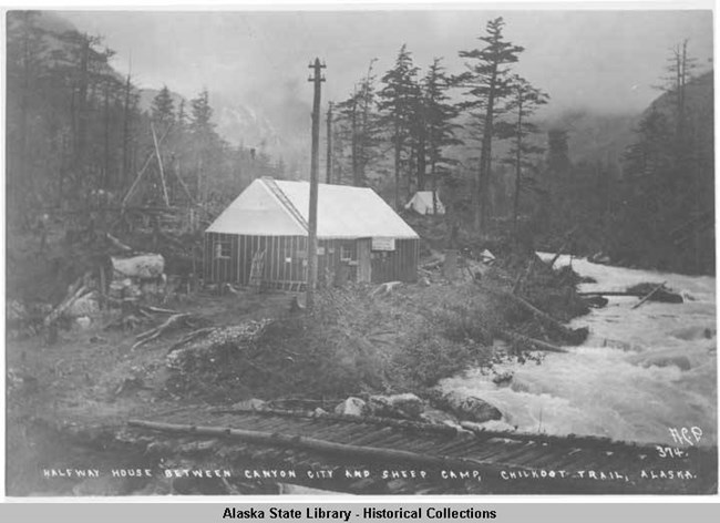 Small cabin, telephone pole, and log bridge next to roaring river in a forested area.