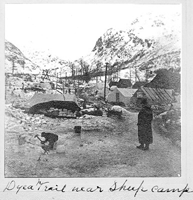 Black and white photo of two people working amid tents and shanties with a snowy mountain backdrop