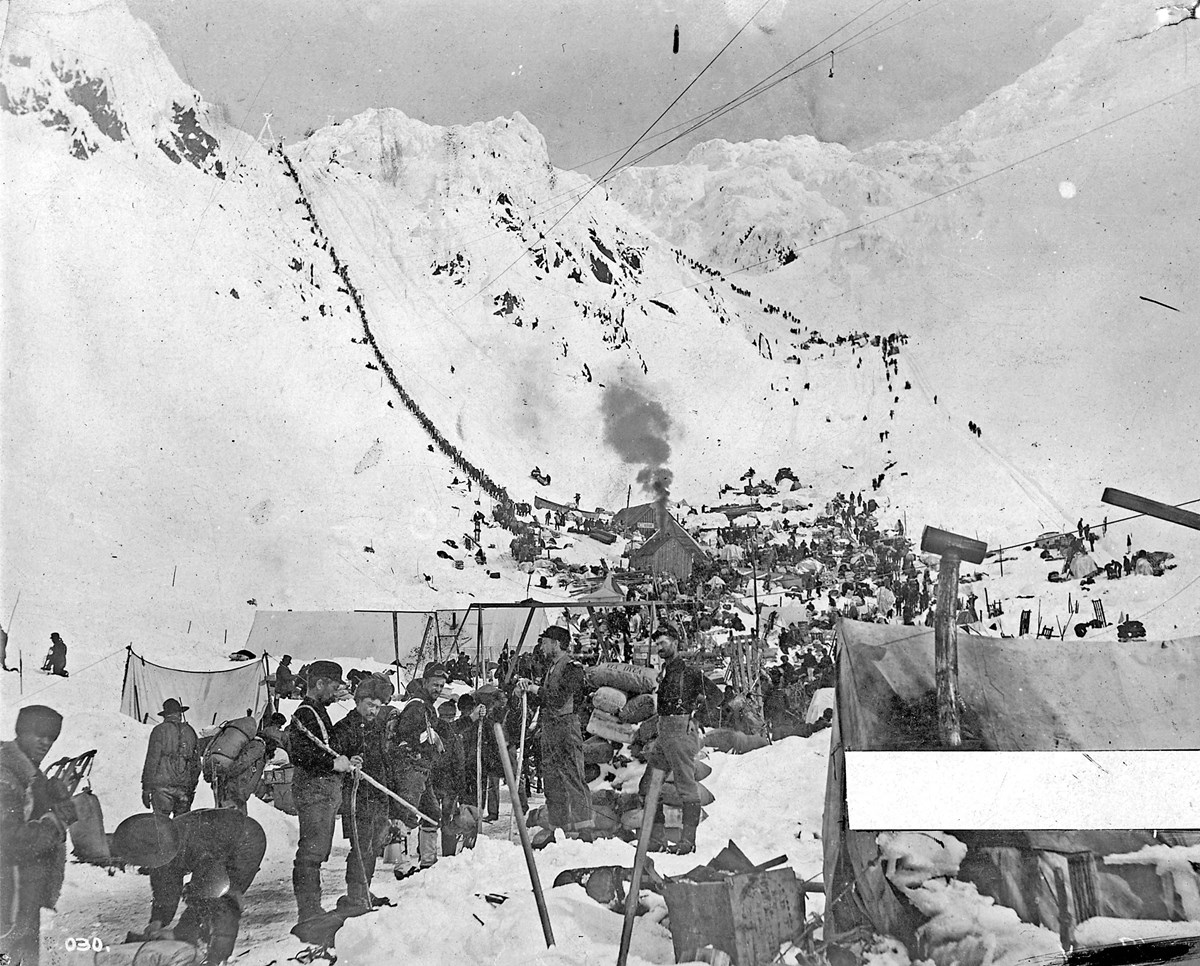 Black and white photo of people and gear and tents gathered at the base of two snowy mountain passes.  Wires cross the sky.