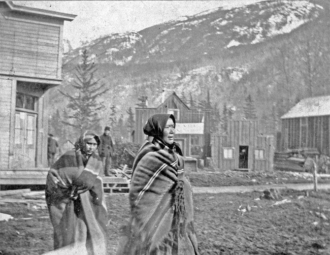 Two Native women wrapped in blankets walk down a muddy street.