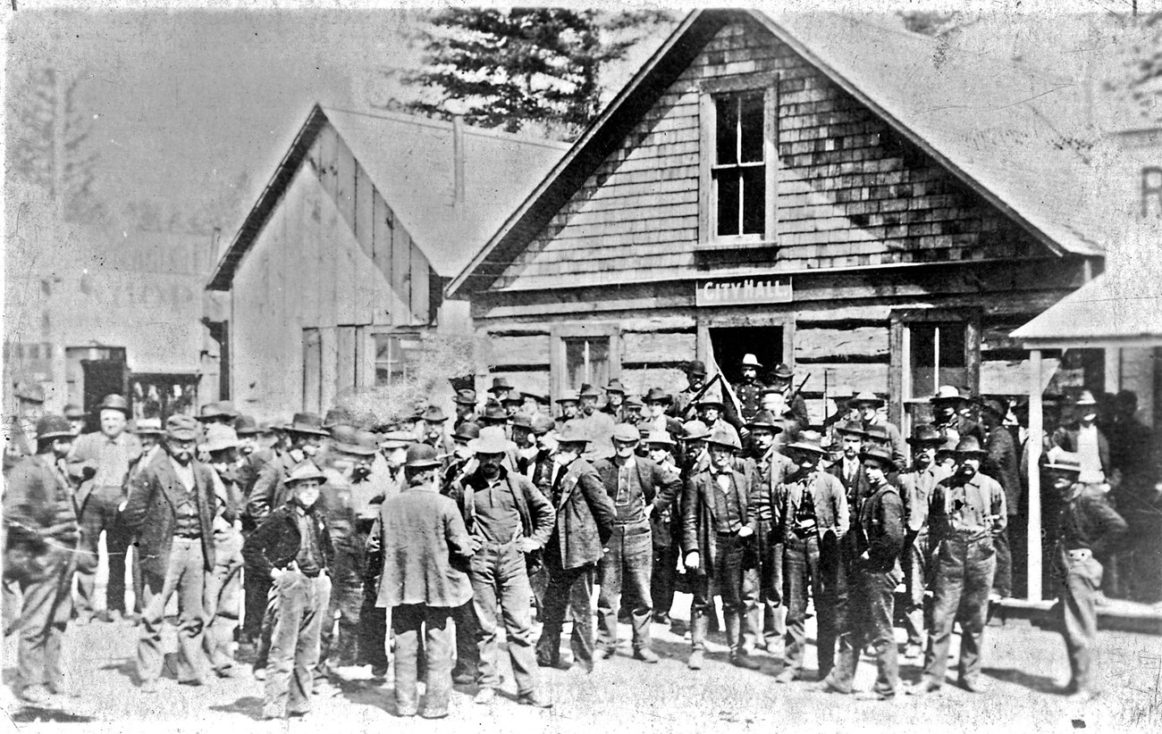 A large group of men gather on the street outside a wooden two story building