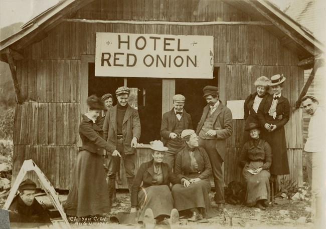 Group of a dozen men and women in old fashioned clothes pose in front of a building with sign "Hotel Red Onion" handwritten caption reads "Canyon City Aug 4th 1901"