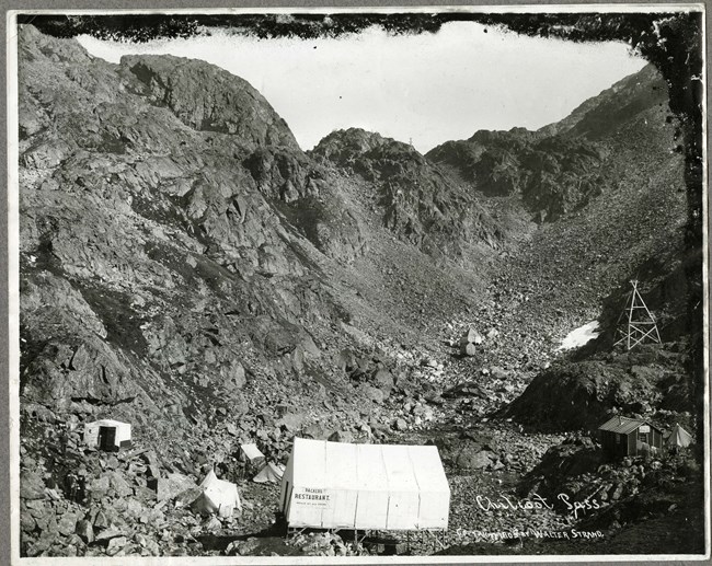 Black and white photo of a few tents and buildings in front of rocky mountain backdrop