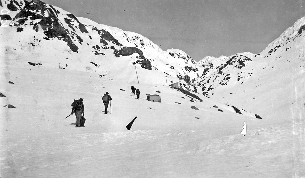 Figures walk up a snowy slope surrounded by snowy mountains.