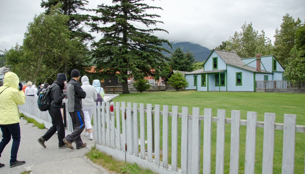 People approach a blue house with a green lawn and picket fence.