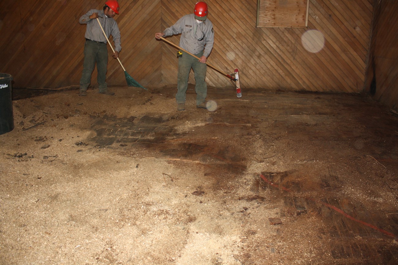 Modern color photograph of two men with a broom and rake clearing the floor of a wooden interior room.