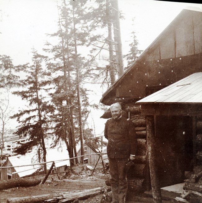 A uniformed man leans against a log cabin