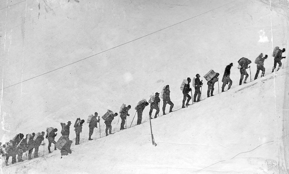 People with gear on their backs stand in a single file line headed uphill with a snowy backdrop.