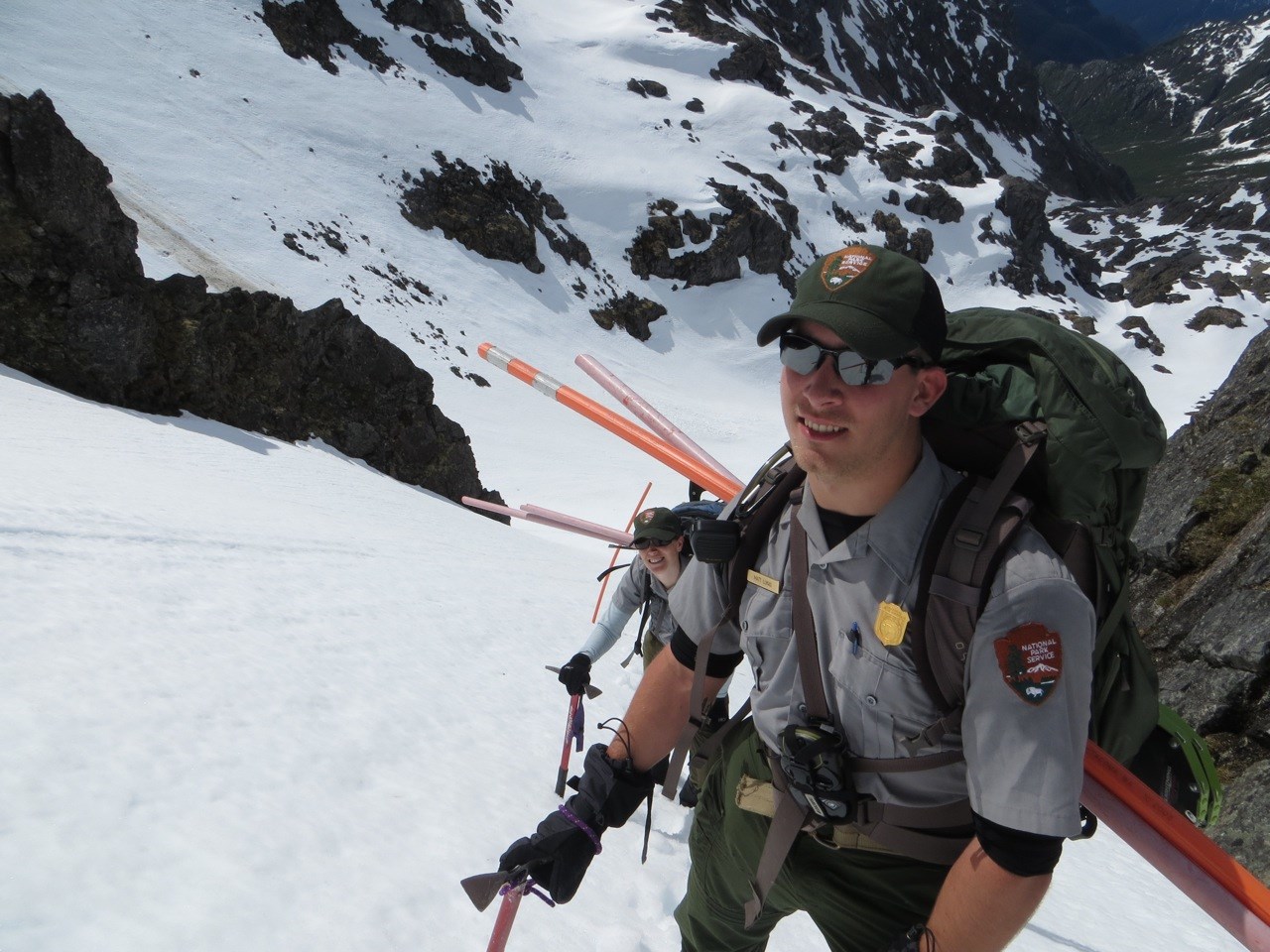 Two rangers carry trail markers up a steep, snowy mountain