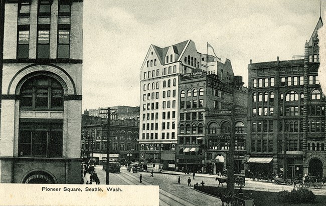 A black and white view of city buildings with a busy street in the middle. Text reads "Pioneer Square Seattle, Wash."