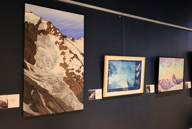 A painting with a brown mountain covered in glaciers, a painting with varying blue tones of a glacier, and a landscape scene of blue, yellow, and purple with glaciers on mountains, hang on a dark blue wall.