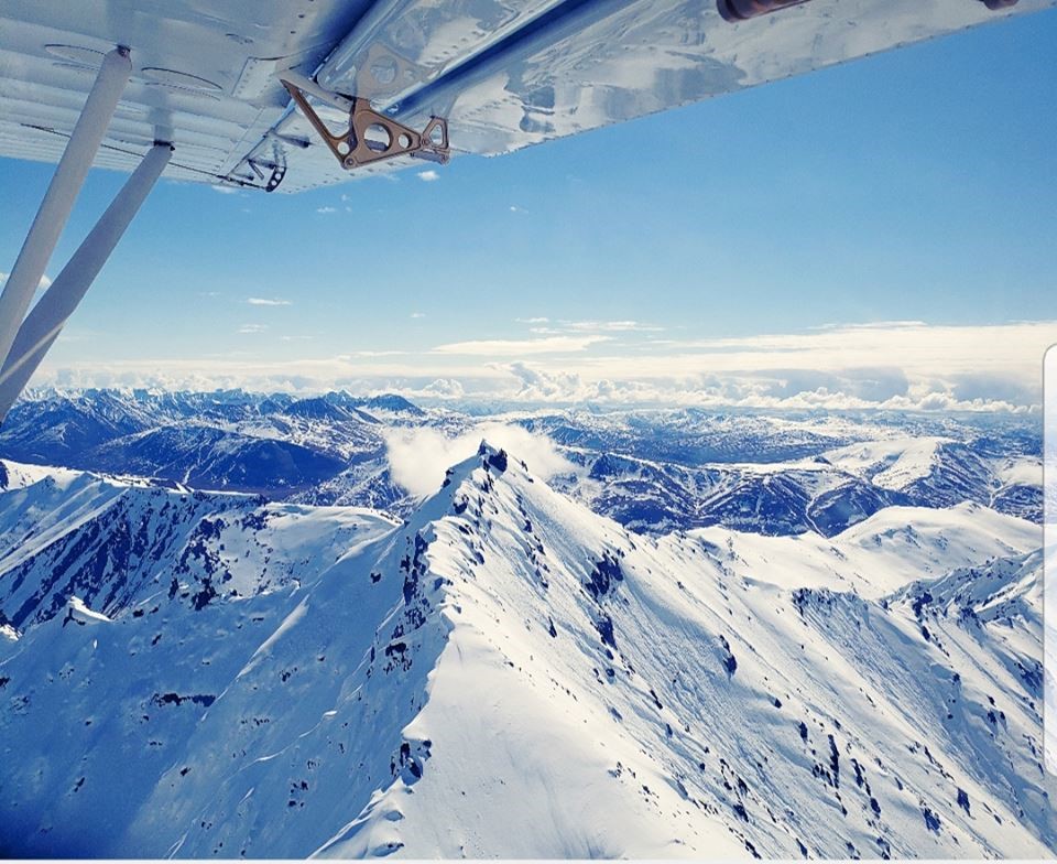 Snowy mountain peaks under a blue sky with bottom of plane wing visible at top