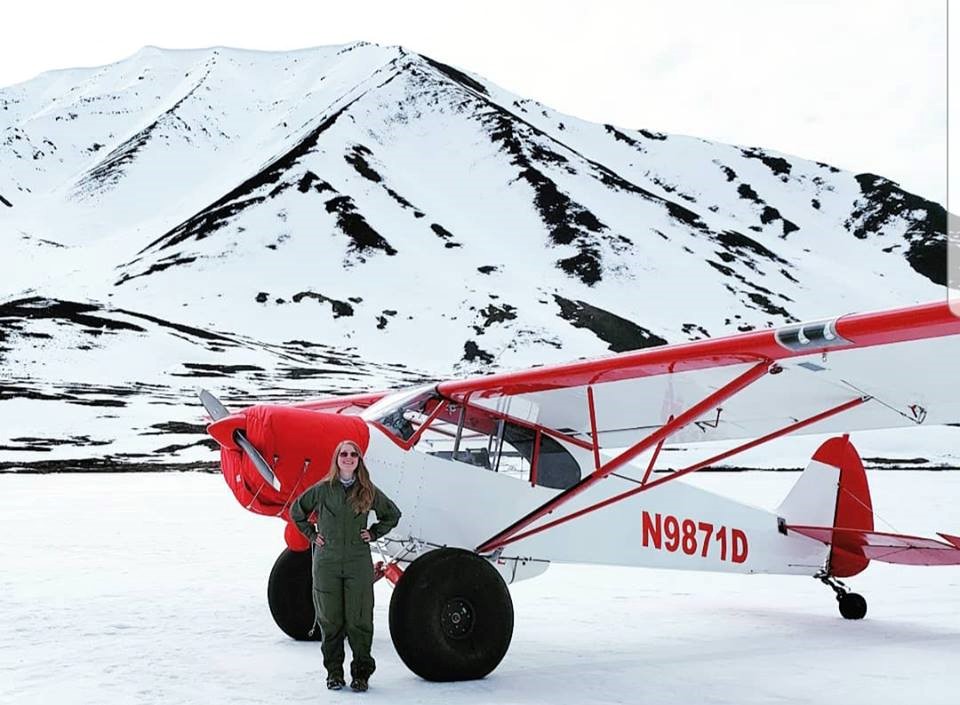 NPS ranger poses in flight suit next to Supercub plane on snowy ground with snowy mountain in background