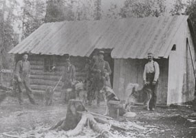 historic image of four men and a few dogs in front of a log cabin with a sheet metal roof