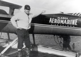 historic, black and white photo of a man standing next to a float plane.