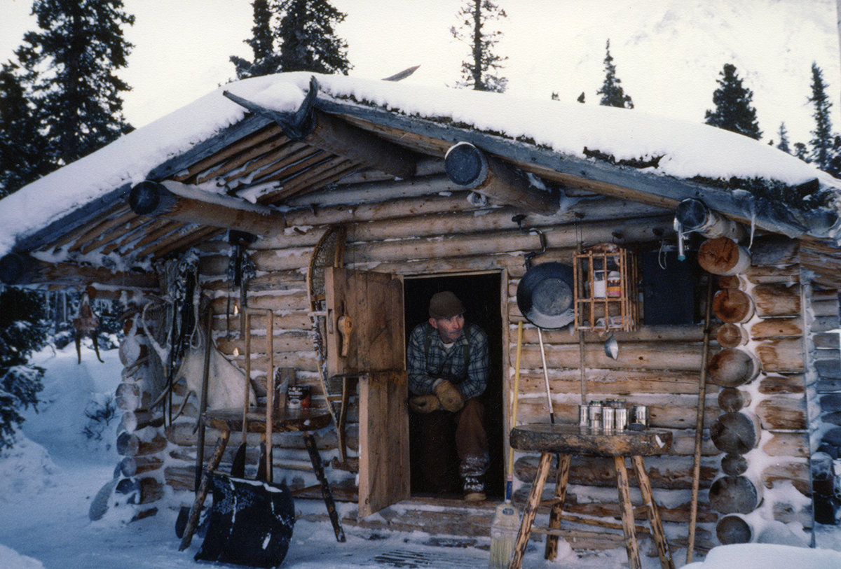 A man wearing a blue flannel shirt and brown pants sits in the open doorway of a log cabin on a winter day.