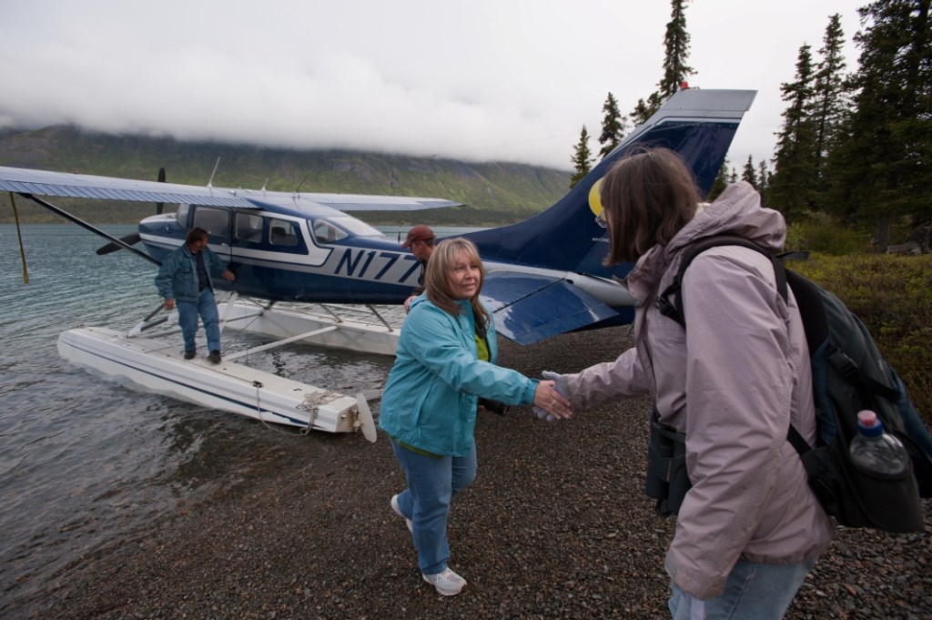 Women shaking hands with blue and white float plane in the background. Two other people are standing on the floats of the plane.