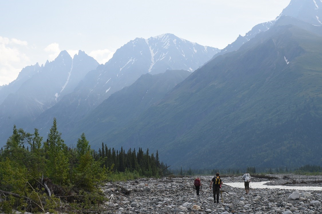 Three hikers walk along a gravel river bed with misty mountains in the background